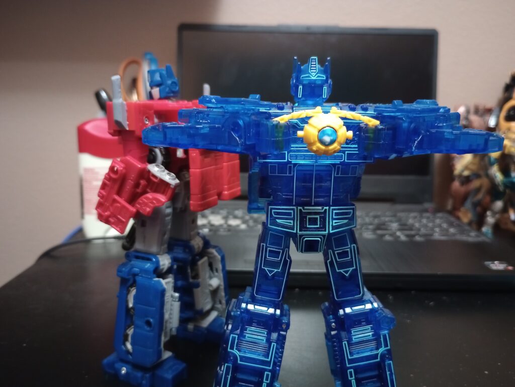 Siege Optimus and Age of the Primes Energon Optimus. The former is facing away from the camera, looking over his shoulder. The latter is standing front and center holding an accessory of the Matrix of Leadership.